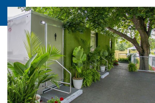 Exterior of a silver luxury restroom trailer decorated with white potted tropical plants and green hedge privacy walls for an upscale outdoor event.