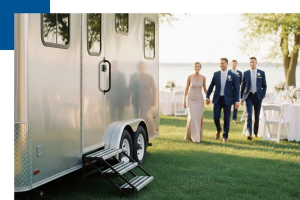 A sleek silver luxury restroom trailer at an outdoor wedding in Big Rapids, with a wedding party walking on the grass nearby.