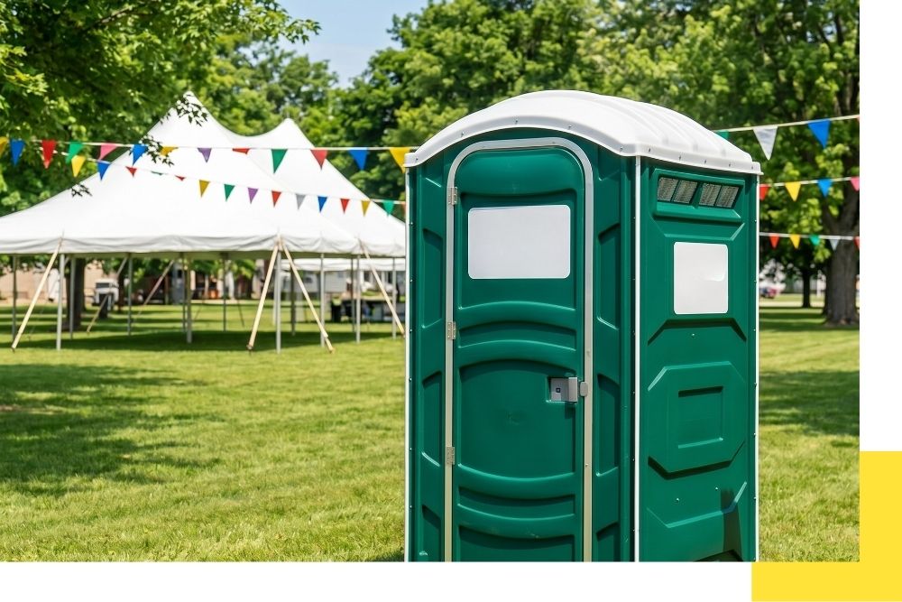 A square stock photo of a clean, deluxe Jons to Go branded porta potty unit set up in a lush park in Bravo, Michigan, with a summer event tent and colorful banners visible in the background.