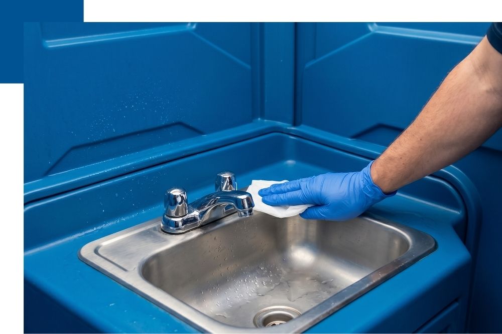 A close-up photograph of a gloved hand performing a final sanitizing wipe-down on the interior stainless steel sink and faucet of a pristine portable restroom unit, emphasizing professional cleanliness.