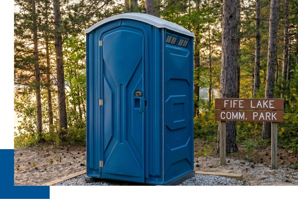 A 1:1 photograph showing a single, unbranded portable restroom unit neatly positioned on gravel in a well-maintained Fife Lake community park setting, near a generic sign that reads 'FIFE LAKE COMM. PARK', surrounded by Michigan white pines.