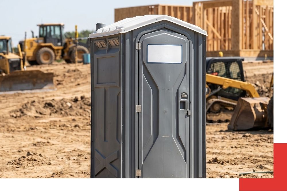 A rugged, grey portable restroom unit with the Millikin Portables logo sits on a dusty construction site near Johannesburg, Michigan, with heavy machinery and building frames blurred in the background