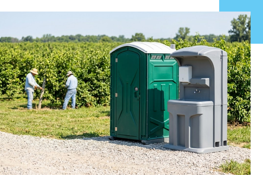 a robust forest green agricultural portable restroom and a separate gray handwashing station. The scene is located near Colfax, Michigan, on a clear compacted gravel path next to an active, lush agricultural field