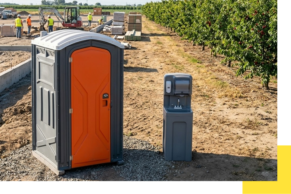 A durable gray plastic portable restroom unit with an orange door and integrated hand sanitizer, alongside a gray handwashing station, placed on gravel at a construction site in Frankfort, MI